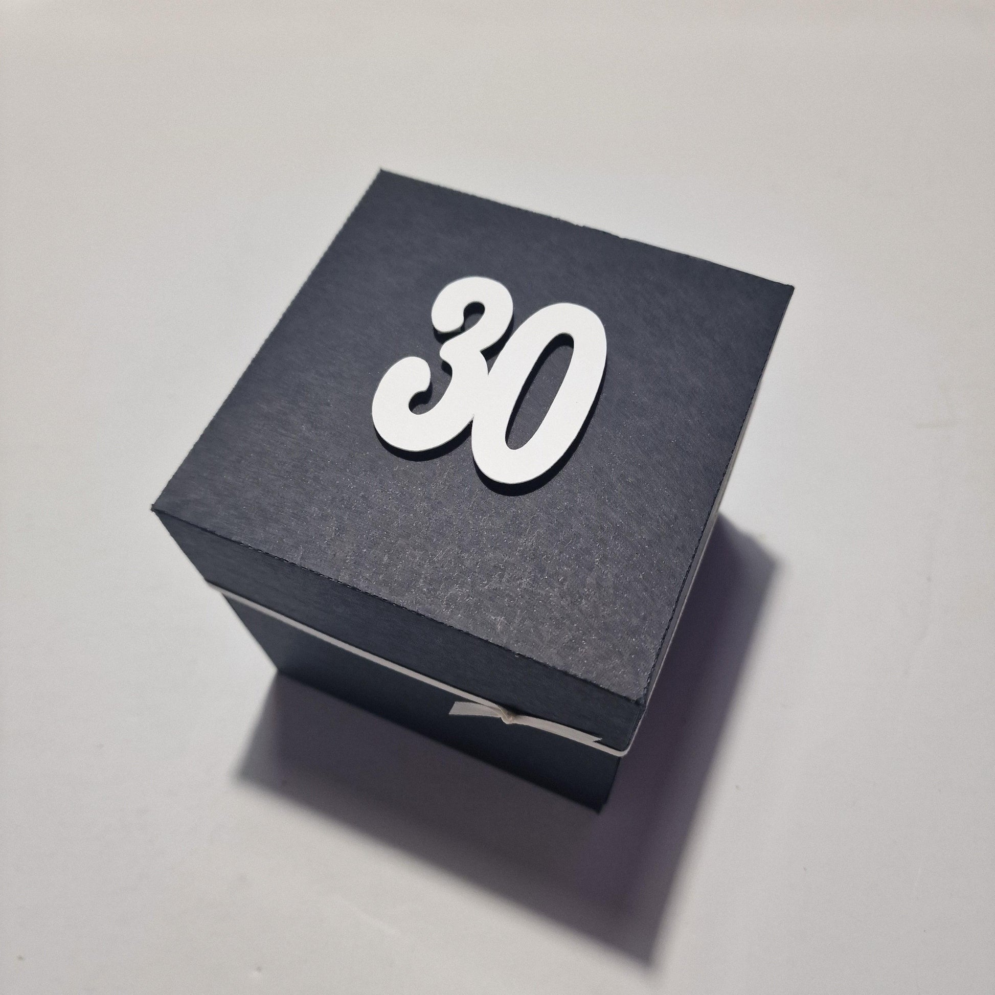 Exploding Occasions Box - Trip Reveal AMSTERDAM: A black box with white numbers and a white ribbon, designed for a surprise Amsterdam trip, featuring iconic canal houses and customizable event details inside.