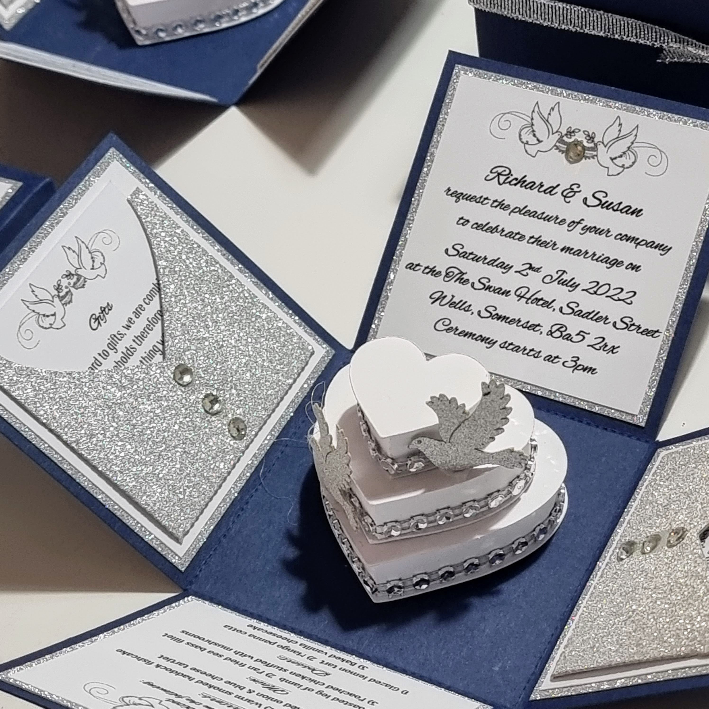 It looks like you're looking at some beautiful wedding invitations! The dark blue boxes with silver ribbons give off such an elegant vibe. And the pop-up card with a three-tiered wedding cake adds a delightful touch! If you're planning a wedding or helping someone with theirs, these are a fantastic choice.