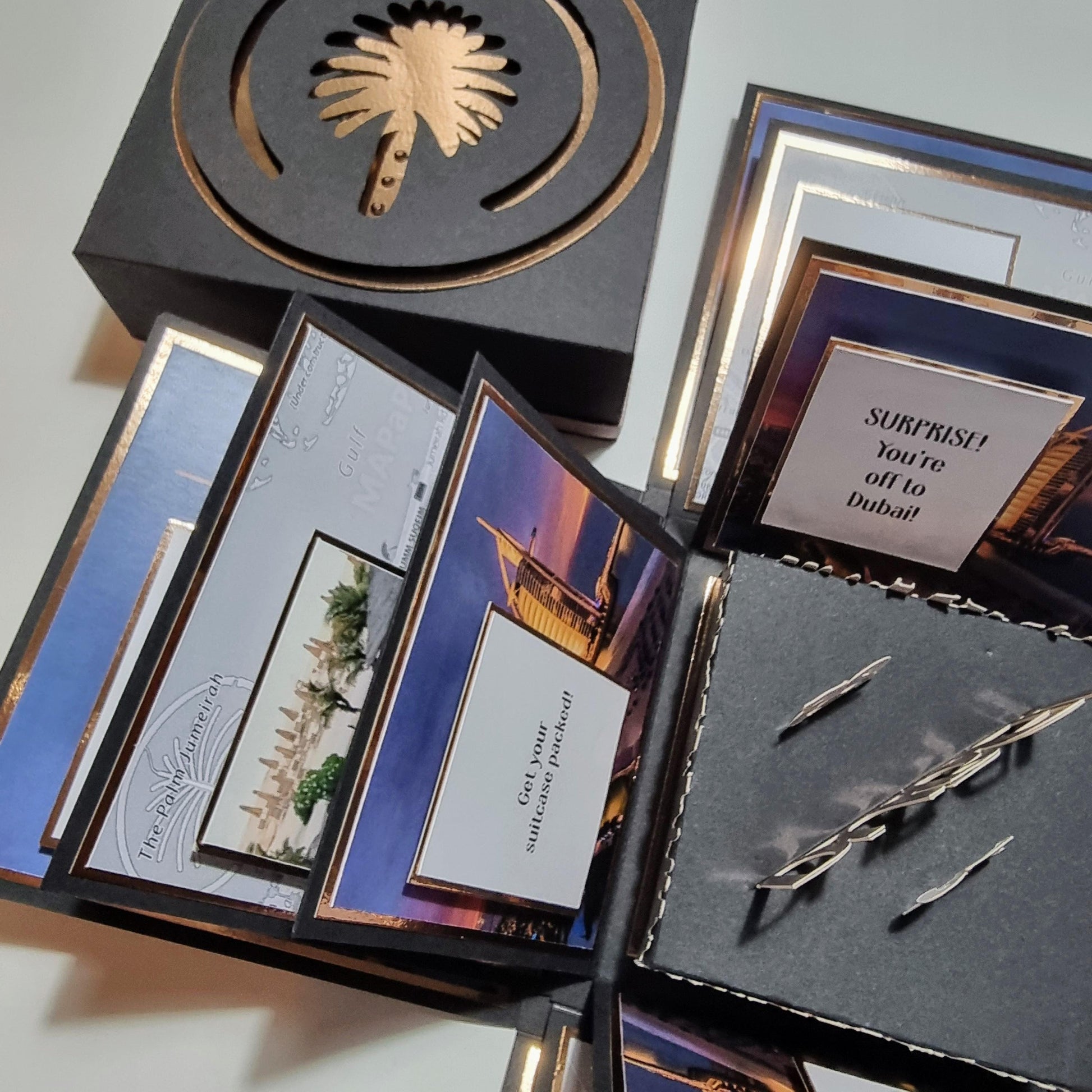 Exploding Occasions Box - Trip Reveal Dubai showcases a black box with gold trim, containing cards and iconic Dubai-themed panels, designed for a surprise travel reveal.