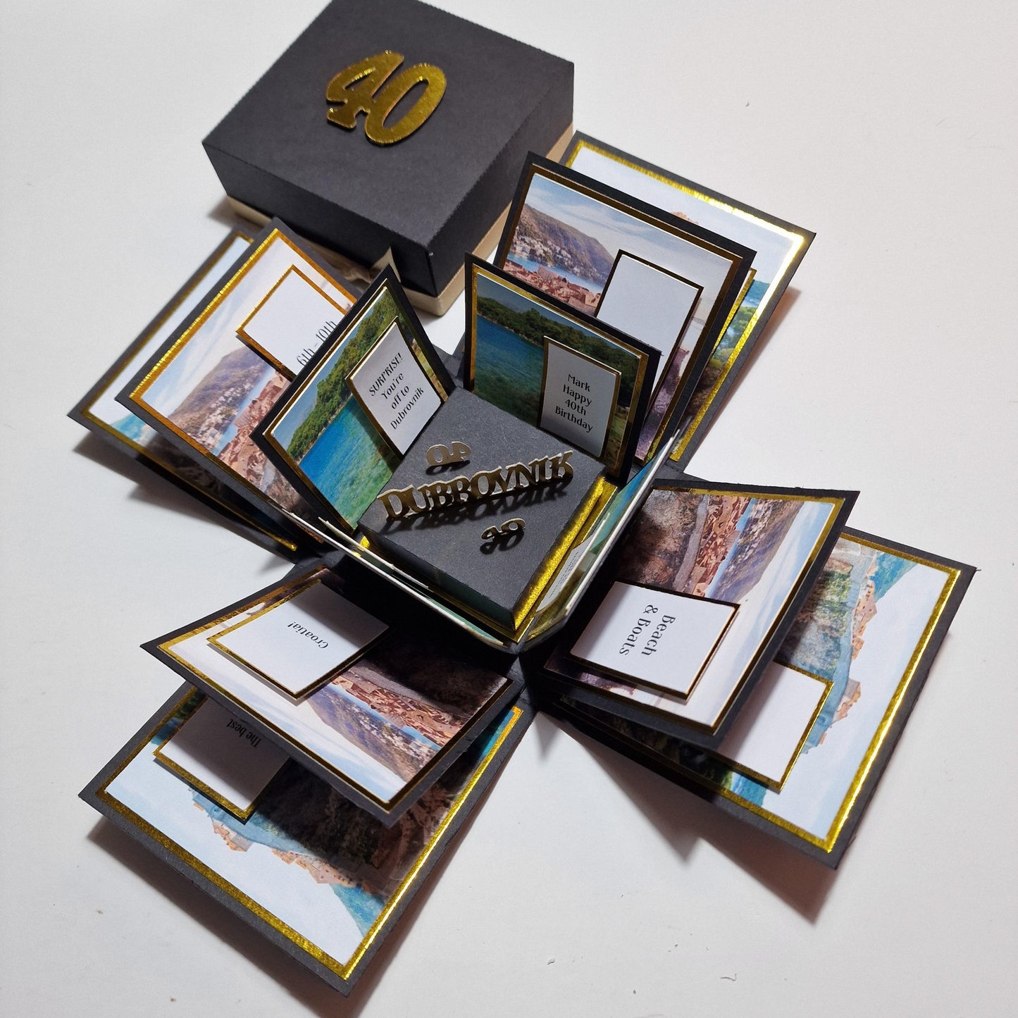 Exploding Trip Reveal Box - DUBROVNIK featuring a black box with gold numbers, containing themed cards and images related to a surprise trip experience.