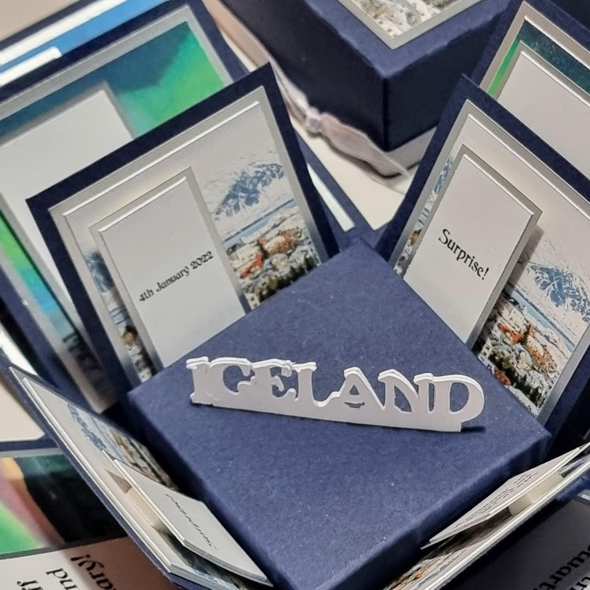 Exploding Occasions Box - Trip Reveal ICELAND featuring cards inside a blue box with a white sign, showcasing Iceland-themed landscape and verse panels for a surprise trip announcement.