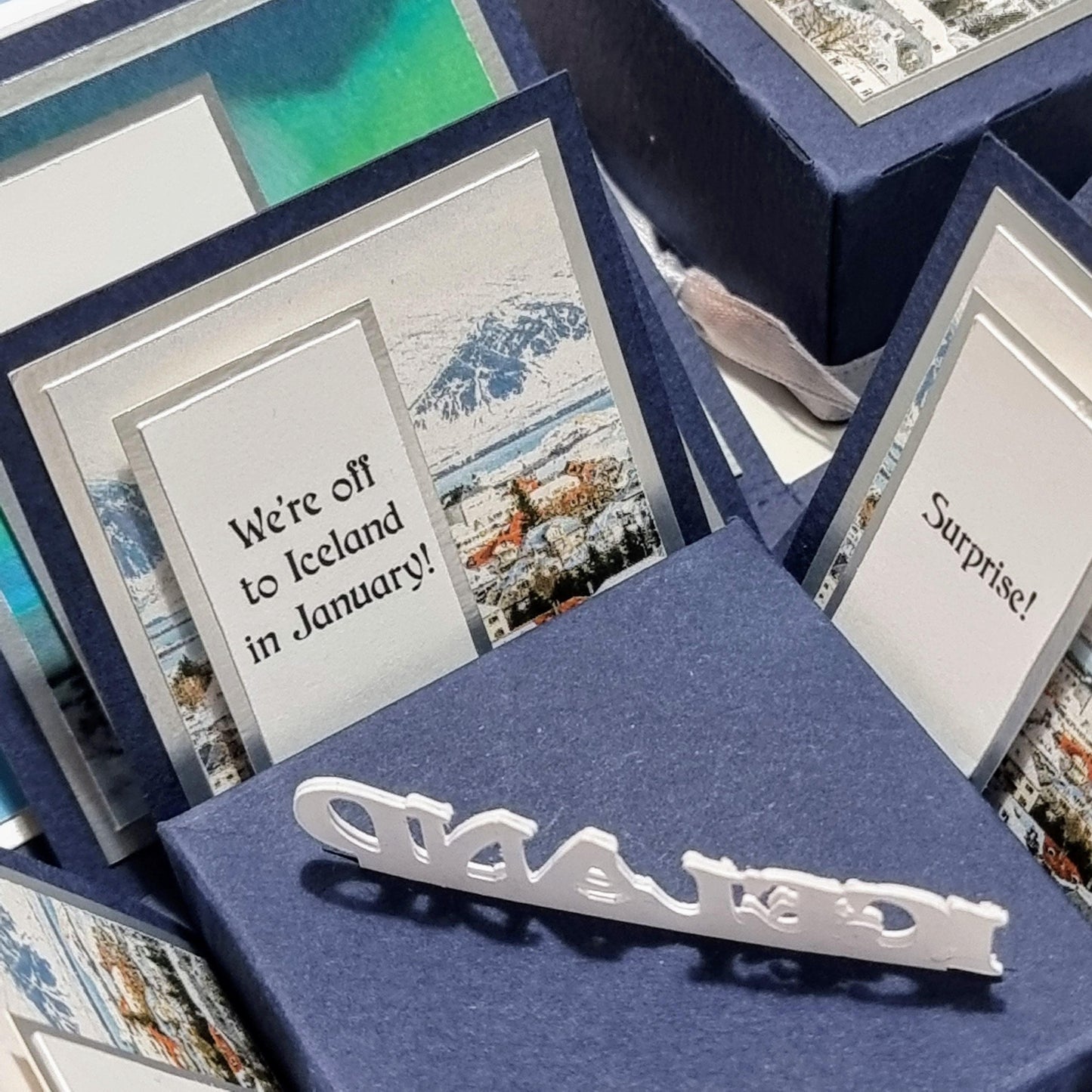 Exploding Occasions Box - Trip Reveal ICELAND: A group of blue boxes featuring Iceland-themed cards, landscape paper panels, and a freestanding Iceland centerpiece for a special surprise trip announcement.