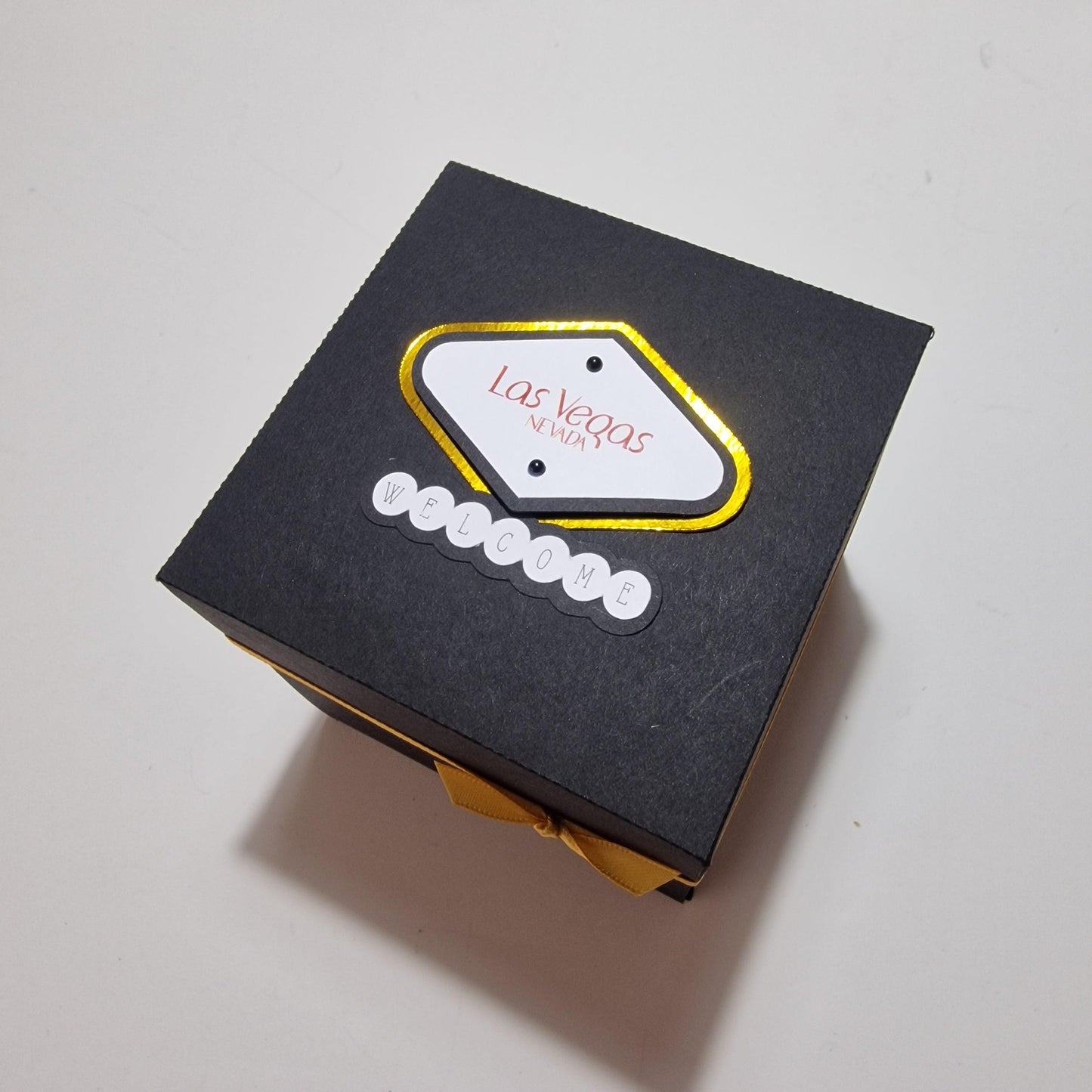 Exploding Occasions Box - Trip Reveal Las Vegas features a black box adorned with a gold ribbon, designed to surprise with Las Vegas-themed elements and a freestanding Las Vegas sign inside.