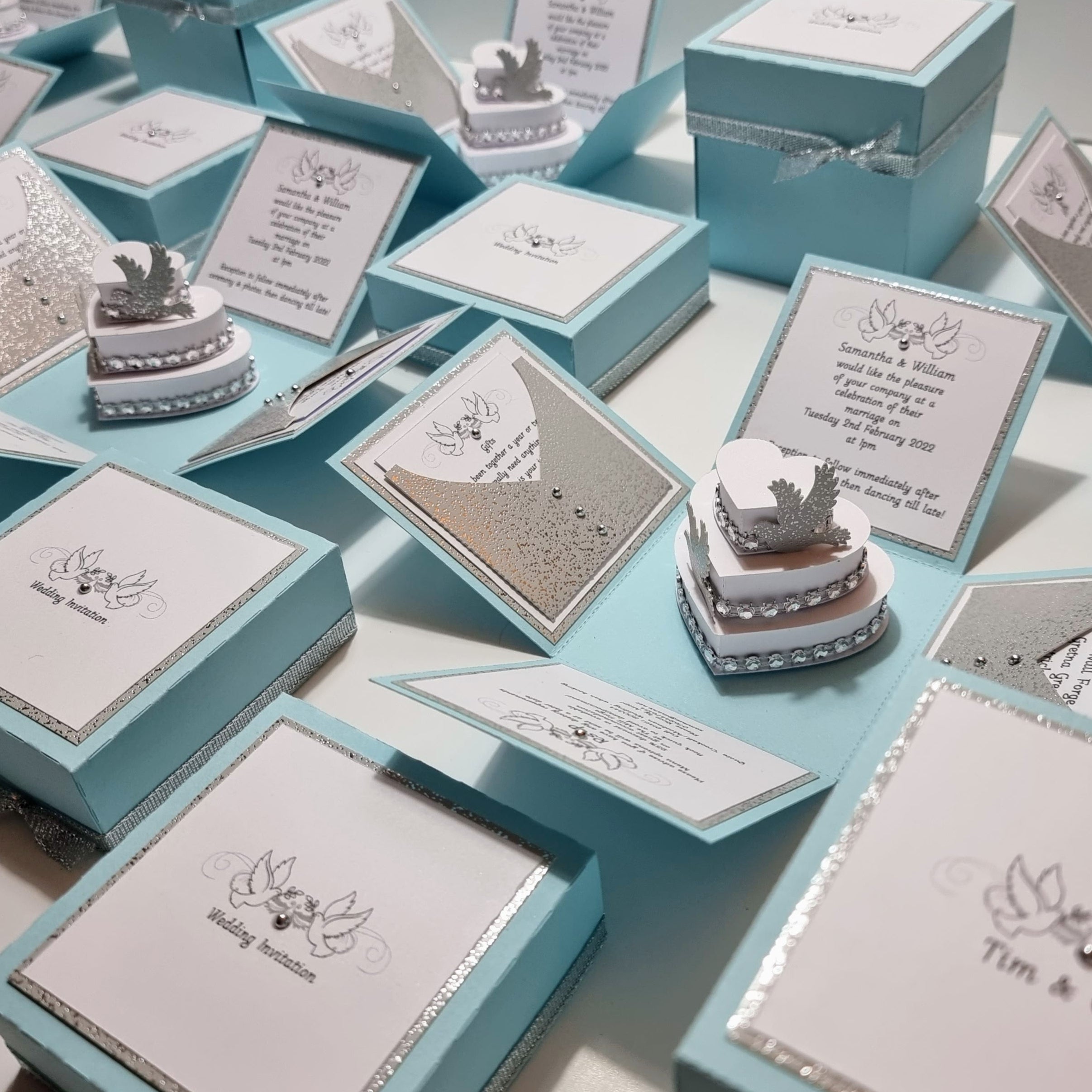 Pastel Blue & Silver Sparkles | Exploding Wedding Invitation Boxes featuring intricate silver designs, fixed panels with invitation and venue details, and a heart-shaped cake topped with doves.