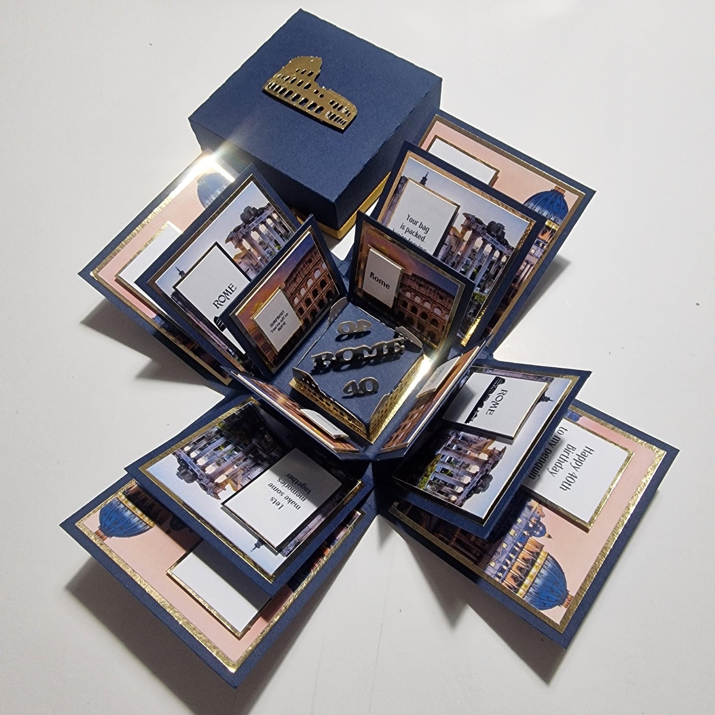 Exploding Occasions Box - Trip Reveal ROME, a blue box featuring Rome's Colosseum image and gold accents, designed for surprise trip announcements with customizable details and themed paper panels.