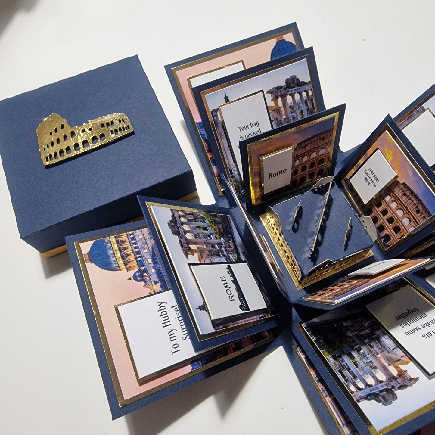 Exploding Occasions Box - Trip Reveal ROME features a blue box adorned with images of Rome, including the Colosseum, and a gold freestanding object, designed for a surprise trip announcement.