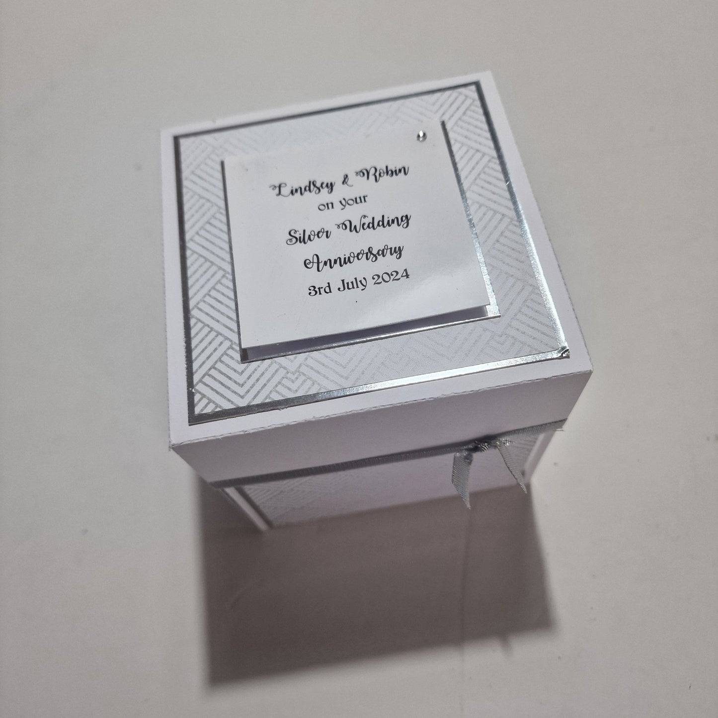 Silver Wedding Anniversary Box featuring a white base, silver ribbon, and a card. Elegant design with customizable panels for names, dates, messages, and photos, celebrating 25 years of love.