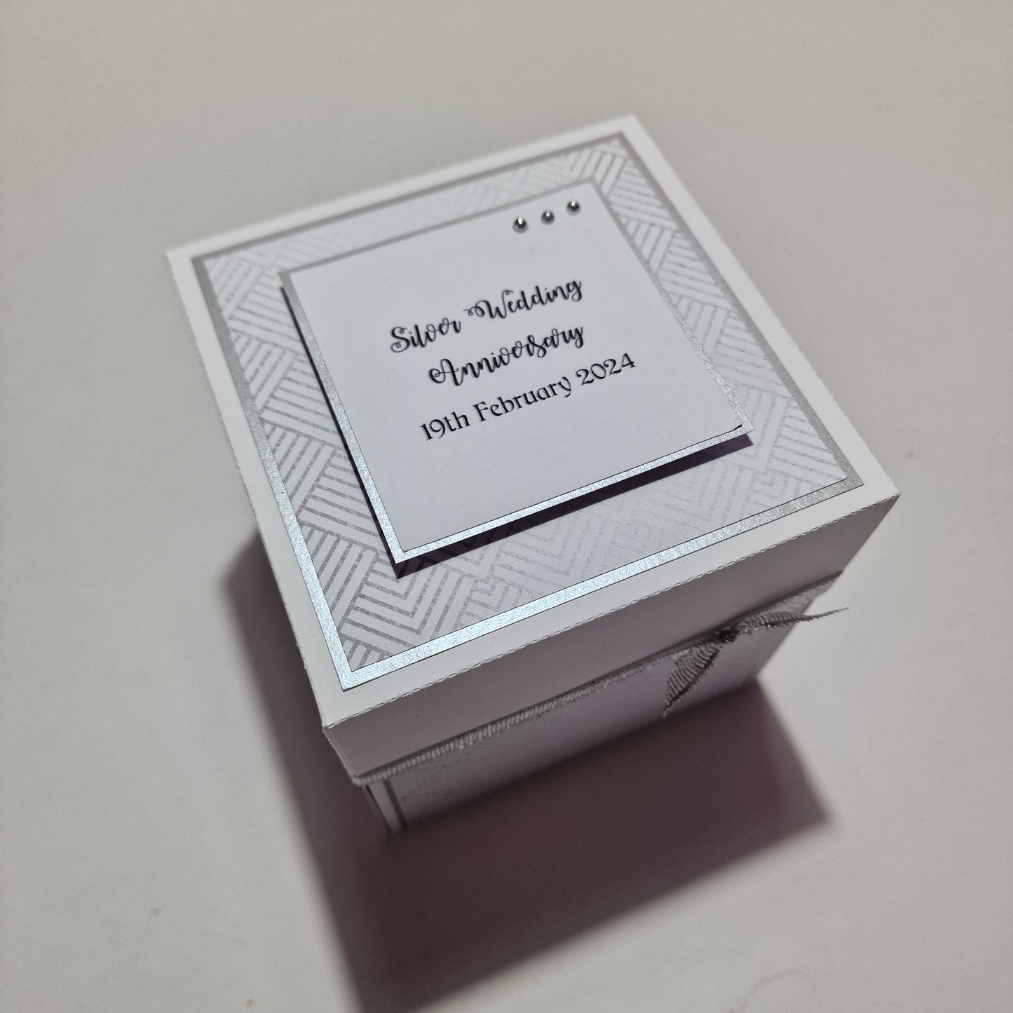 Silver Wedding Anniversary Box featuring a white base with silver ribbon and card, designed to celebrate 25th anniversaries. Elegant keepsake for personal messages and photos.