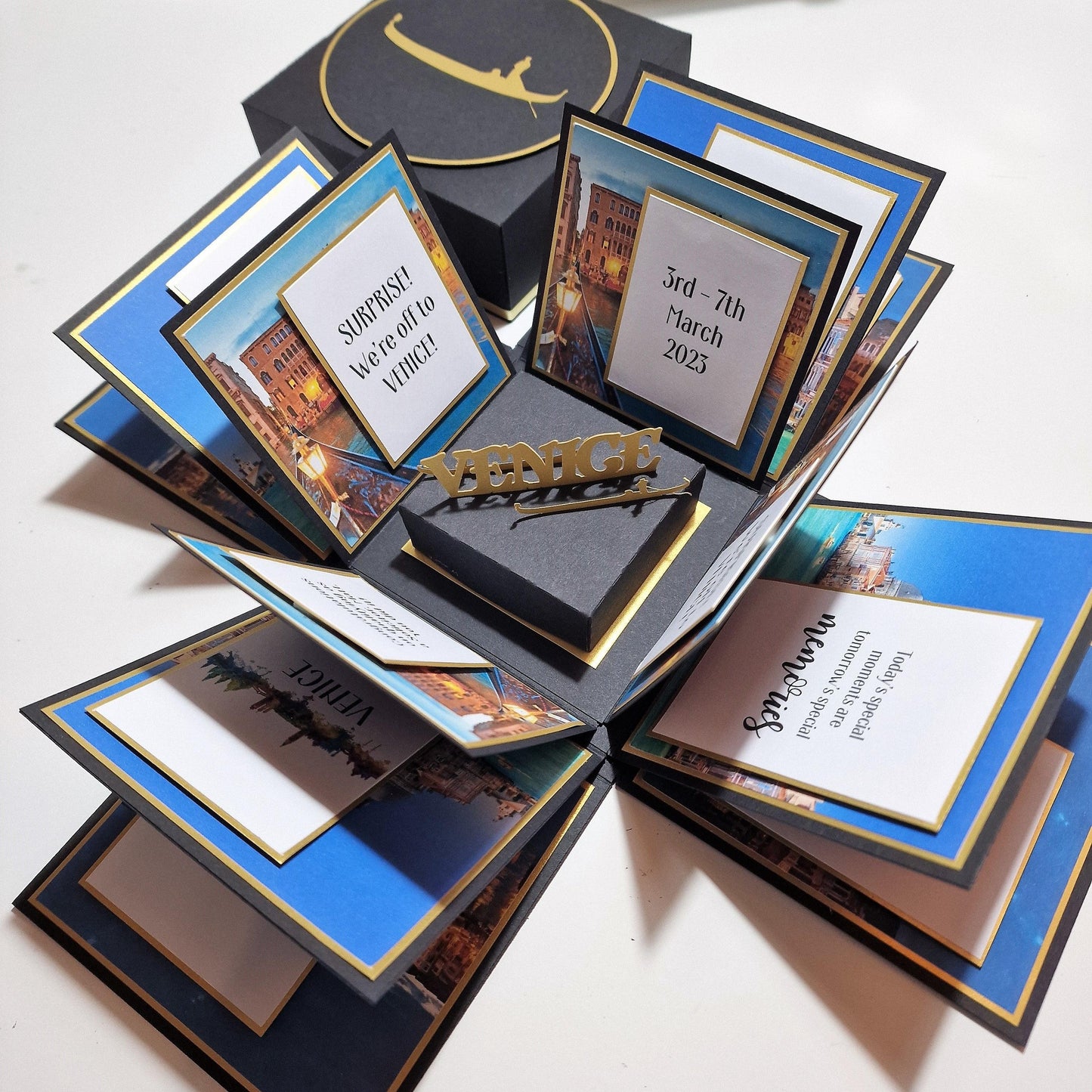 Exploding Occasions Box - Trip Reveal VENICE: A black box with gold lettering, containing themed Venice trip elements, including a gondola and iconic architecture panels, with personalized card details inside.