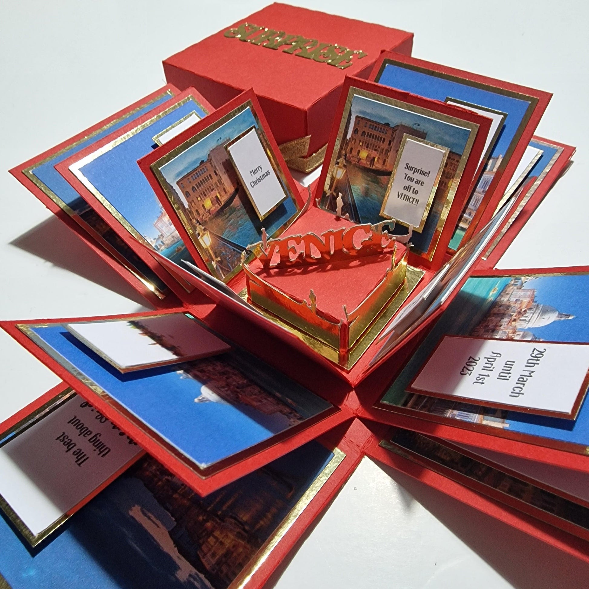 Exploding Occasions Box - Trip Reveal VENICE features a red box adorned with gold letters and a ribbon, showcasing Venice-themed panels, iconic architecture, and a gondola centerpiece.