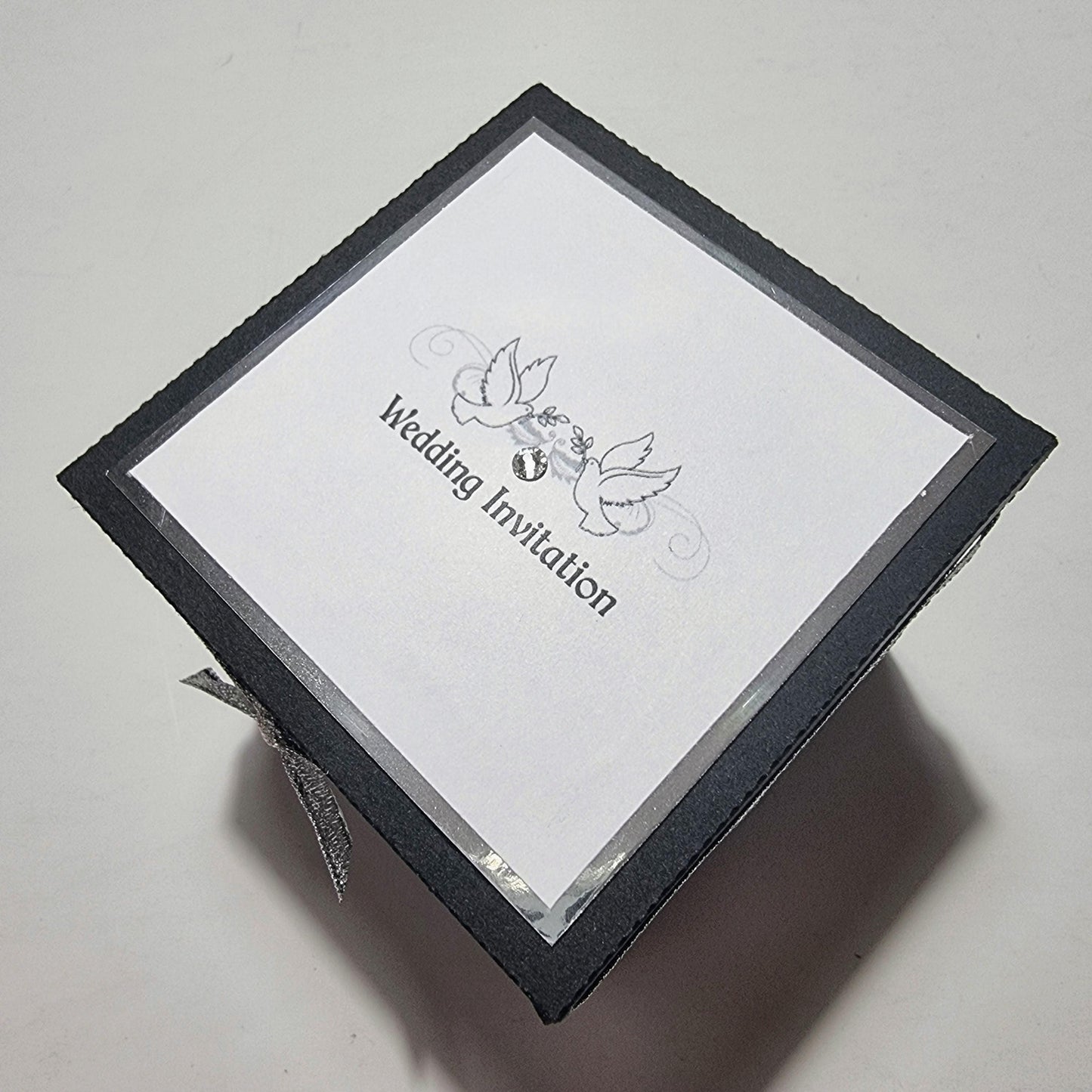 Black & Silver Exploding Wedding Invitation Box with dove-decorated tiers, featuring fixed invitation and venue panels, waistcoat pockets with pull-out menu, RSVP cards, and a heart-shaped cake centerpiece.