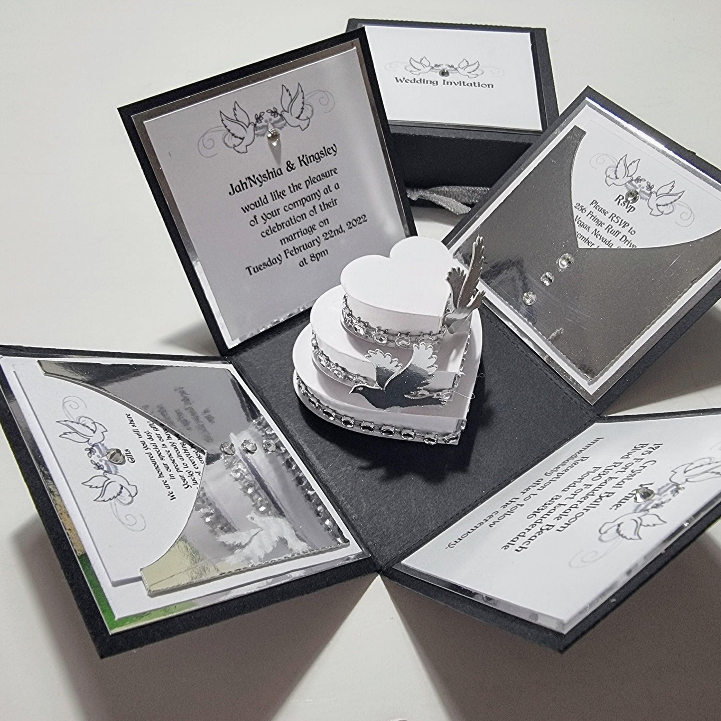 Black & Silver Exploding Wedding Invitation Box featuring heart-shaped compartments, a central tiered cake with doves, and elegant invitation details. Part of the Hearts & Doves Range in Classic Black & Silver.